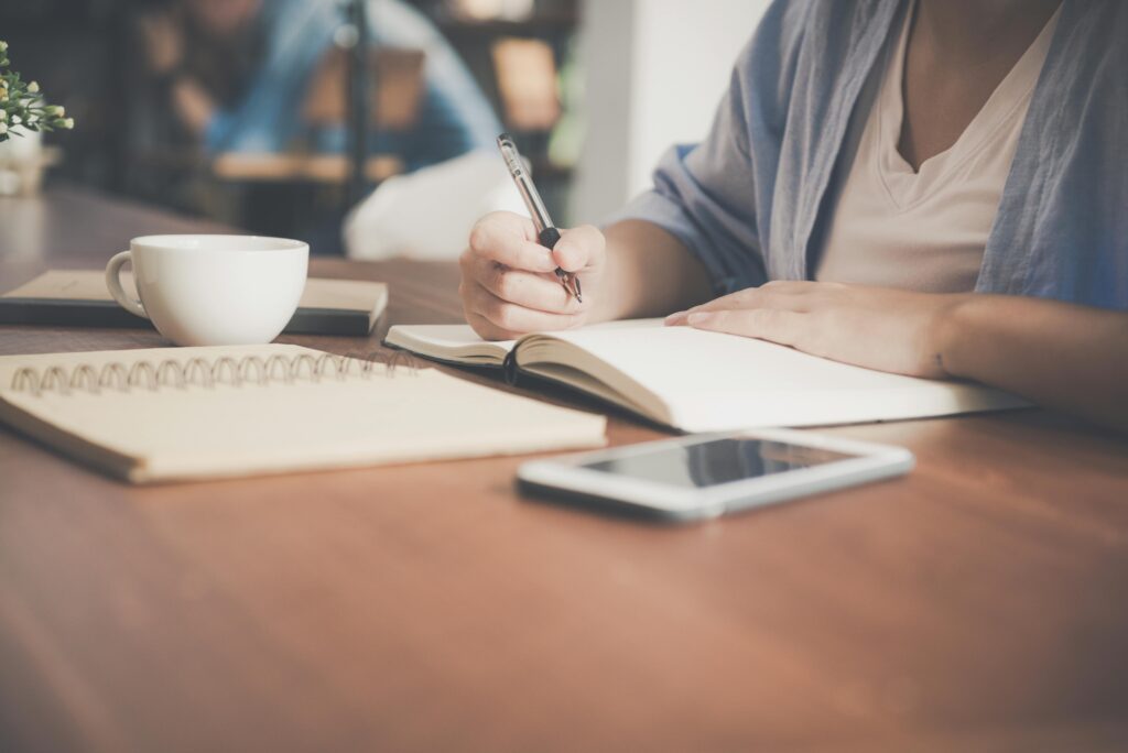 A woman wear a white tshirt and blue cardigan sits at a desk with a cup, three notebooks, and a mobile phone. She is writing in one of the notebooks with a ball pen.